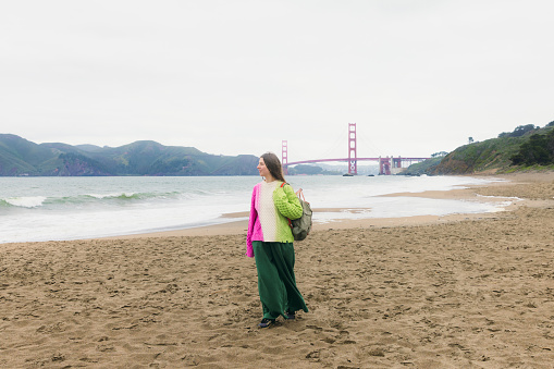 Front view of woman with long hair, in colorful knitted sweater walking at the scenic beach and enjoying music from headphones with background view of red Golden Fate Bridge in San Francisco city, the United States