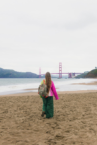 Rear view of woman with long hair, in colorful knitted sweater walking at the scenic beach and enjoying music from headphones with background view of red Golden Fate Bridge in San Francisco city, the United States