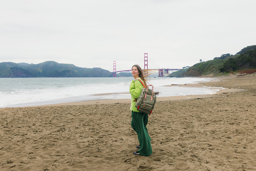 Side view of woman with long hair, in colorful knitted sweater walking at the scenic beach and enjoying music from headphones with background view of red Golden Fate Bridge in San Francisco city, the United States