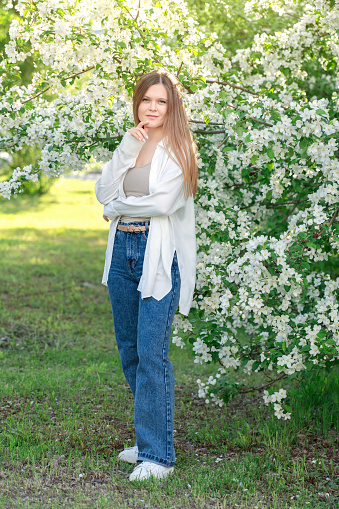 One blond young woman wearing white blouse and blue casual jeans looking straight at camera in the garden in front of blooming apple trees with small flowers and green leaves standing on grass lawn