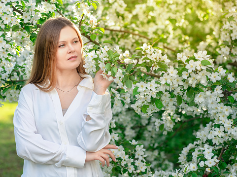 Portrait of blond young woman wearing casual white blouse or shirt in the garden among blooming apple trees with small flowers and green leaves standing with thoughtful smile at sunlight in spring