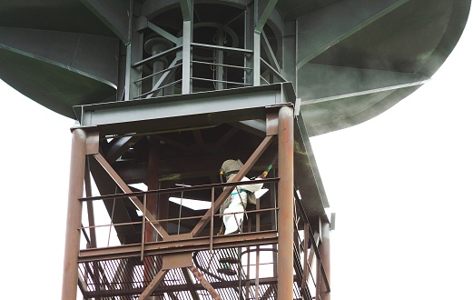 Sand blasting process. Industrial worker in protective uniform cleaning surface of big steel construction before painting on high altitude.