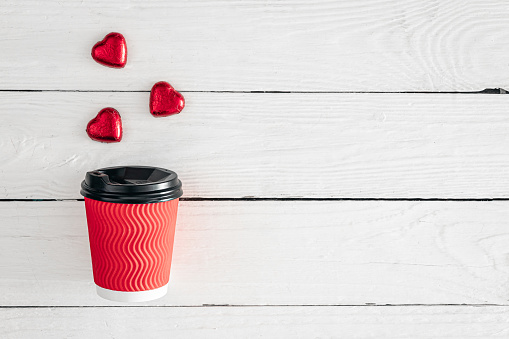 Red paper cup and heart-shaped candies on a white wooden background. View from above. Space for text. Valentine's Day concept.