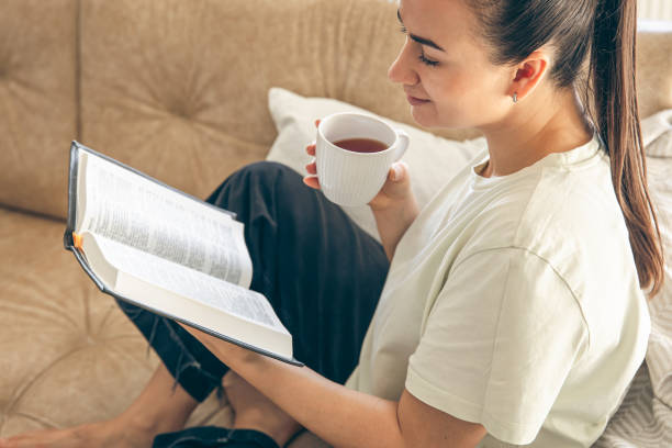 istock Young woman drinking tea and reading book sitting on sofa at home. 1885166308