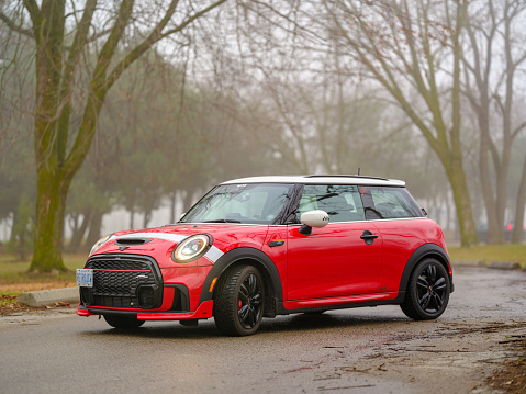 Toronto, Ontario, Canada- December 24, 2023.  Chili red colour MINI COOPER in public park in Toronto East side, Canada. Foggy morning with overcast sky.  This is the third generation model F56 JCW, since BMW took over iconic brand of MINI. MINI featured in the photo is John Cooper Works model, the most powerful 2 door version. For the first time, this compact car features engine build and designed by BMW, and packs even more power and torque than previous models since 2002 to present. Original design clues and themes are still present on this brand new model. Mini has been around since 1959 and has been owned and issued by various car manufacturers.