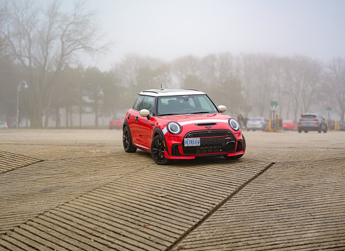 Toronto, Ontario, Canada- December 24, 2023.  Chili red colour MINI COOPER in public park in Toronto East side, Canada. Foggy morning with overcast sky.  This is the third generation model F56 JCW, since BMW took over iconic brand of MINI. MINI featured in the photo is John Cooper Works model, the most powerful 2 door version. For the first time, this compact car features engine build and designed by BMW, and packs even more power and torque than previous models since 2002 to present. Original design clues and themes are still present on this brand new model. Mini has been around since 1959 and has been owned and issued by various car manufacturers.