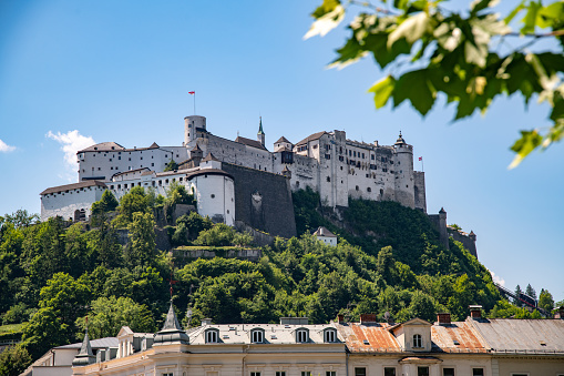Salzburg, Salzburg - Austria - 06-17-2021: View of Hohensalzburg Fortress with houses and trees in the foreground under a graceful, large fortress against a blue sky with light clouds.
