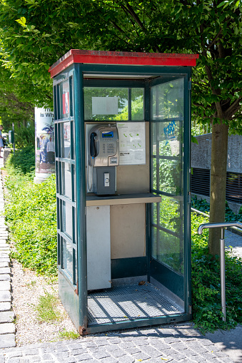 Salzburg, Salzburg - Austria - 06-17-2021: Green telephone booth with red roof, made of glass and metal, without a door. Inside, a telephone and a shelf are visible. Shining sun.