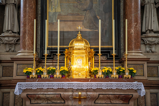 Salzburg, Salzburg - Austria - 06-17-2021: Small altar in Salzburg Cathedral with a golden shrine, surrounded by candles, yellow flowers, a painting, and columns.