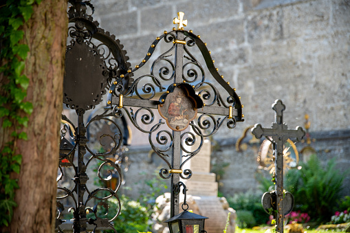 Salzburg, Salzburg - Austria - 06-17-2021: A black metal tombstone or grave cross adorned with many metal decorations. On the grave cross, there is a painting of Mother Mary with the baby Jesus. In the background, a wall and other tombstones can be seen.