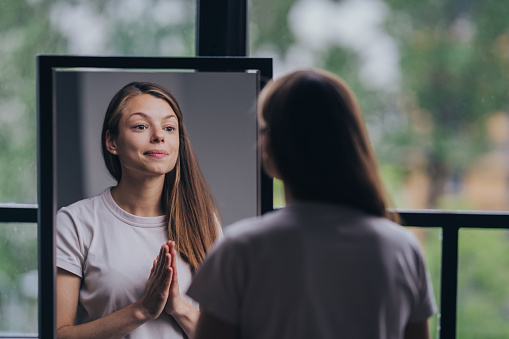 Reflective young woman in a casual t-shirt practicing self-affirmation in front of a mirror in a well-lit room.