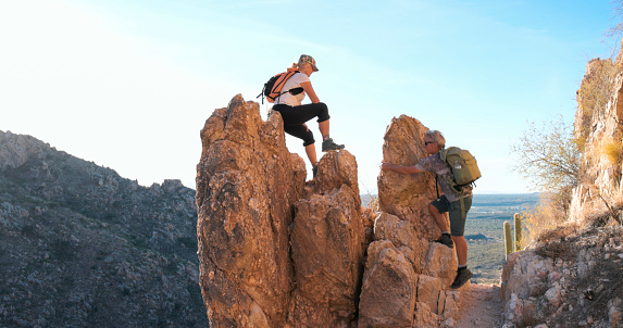 Mature woman balances on rock pinnacles above desert landscape and partner scrambles up to meet her