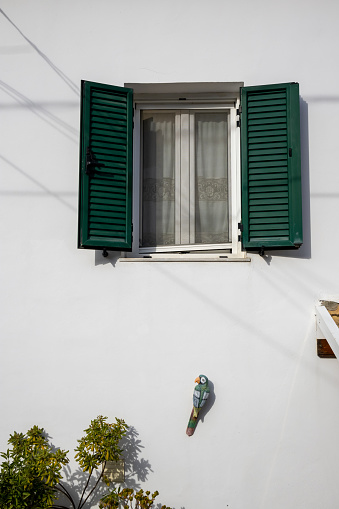 Detail of the White house, where was living british writer Lawrence Durrell. Window with green opened shutters. Wooden parrot decorating the wall. Kalami, Corfu, Greece.