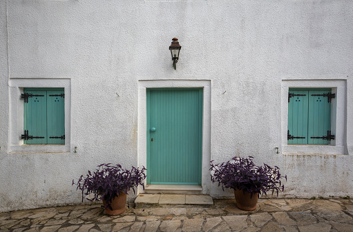 White facade of a house with bright green closed wooden door and window shutters. Lantern above the door. Two pots with violet plants on the stone pathway. Kalami, Corfu, Greece.