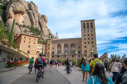 Montserrat, Barcelona Province, Spain - 10-29-2021: A road leads to Montserrat Monastery with many visitors. The buildings stand before a mountain and a blue sky. The paths are very clean.