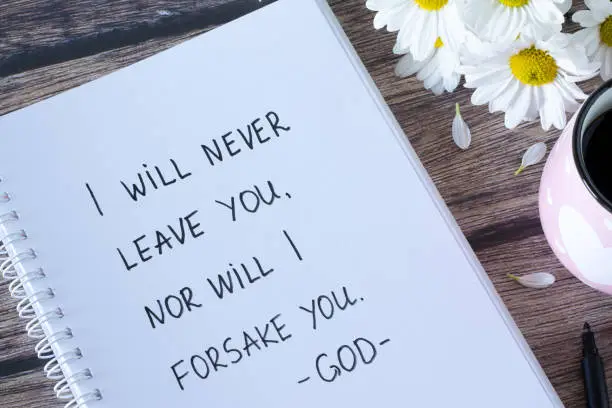 I will never leave you nor will I forsake you-God, handwritten verse in notebook with flowers and coffee cup on wooden table. Book of Hebrews 13:5 text. Top view.