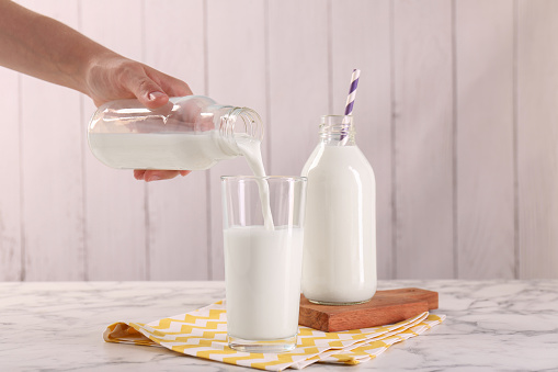 Woman pouring milk from bottle into glass at white marble table, closeup