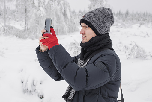 A young handsome caucasian guy in winter clothes takes pictures on a smartphone of a landscape in a winter forest reserve in Belgium, close-up side view. The concept of winter holidays,using technologies.