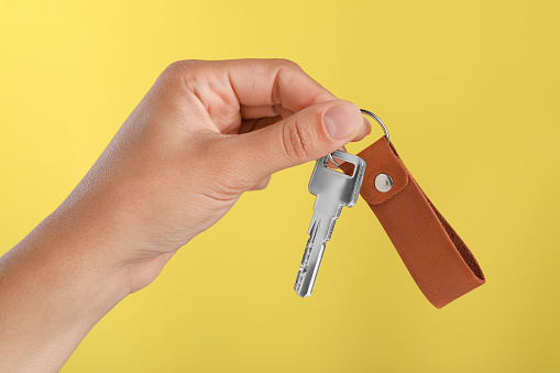 Woman holding key with leather keychain on yellow background, closeup