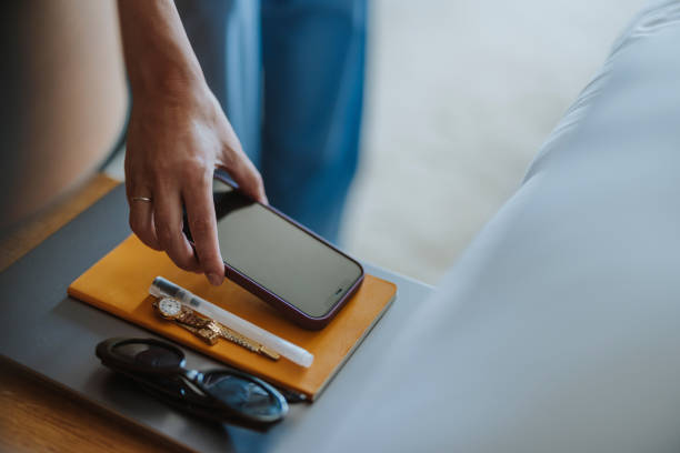 Close up shot of an unrecognizable woman picking up a smartphone from a bedside table in the hotel. There are sunglasses, notebook, watch and pen on the table.