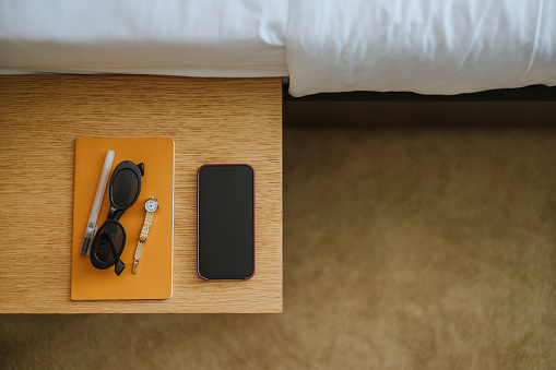 Flat lay of a bedside table in the hotel. There is a mobile phone, notebook, pen, watch and sunglasses.