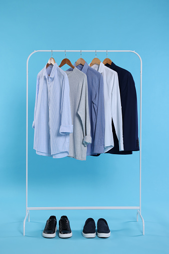Rack with stylish clothes on wooden hangers and sneakers against light blue background