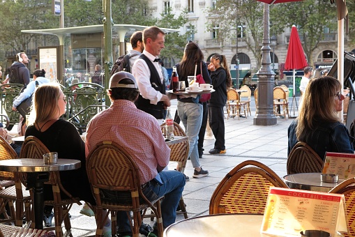 Paris, France - Oct. 3, 2023:  A waiter wearing a black bow tie, vest and white apron hurries to a table with drinks at a sidewalk cafe at the corner of Boulevard and Quai Saint Michel, on the Left Bank of the Seine River.