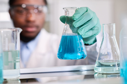Close up shot of conical flask with blue liquid substance in hand of defocused African American laboratory specialist putting container on shelf