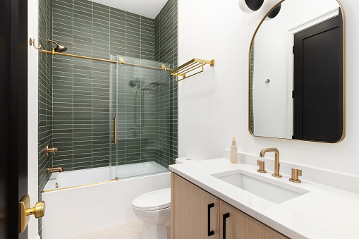 A bathroom with a wood vanity cabinet, gold hardware and faucet, and a shower with green subway tiles.