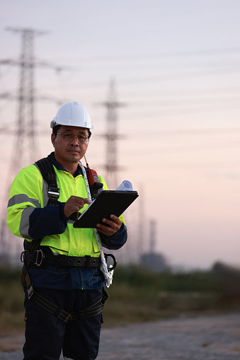 Electrical engineer with green reflec safety jacket and safety harness using tablet and blue print drawing working at site line of electrical distribution