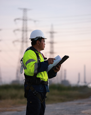 Senior engineer with green reflec safety jacket and safety harness work on tablet and blueprint drawing  at site line of electrical distribution