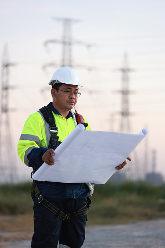 Senior Electrical engineer with green reflec safety jacket and safety harness work on blueprint drawing  at site line of electrical distribution