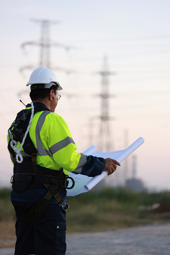 Senior Electrical engineer with green reflec safety jacket and safety harness work on blueprint drawing  at site line of electrical distribution