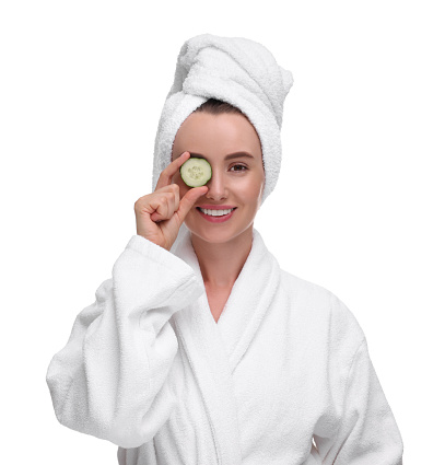 Beautiful woman in bathrobe covering eye with piece of cucumber on white background
