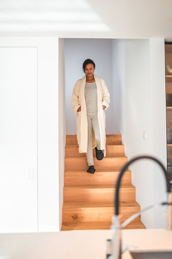 Full length shot of a mixed race female walking down the stairs. She is wearing pijamas and a bathrobe. She is sleepy.