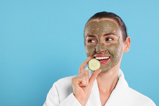 Beautiful woman with clay mask holding piece of cucumber on light blue background, space for text