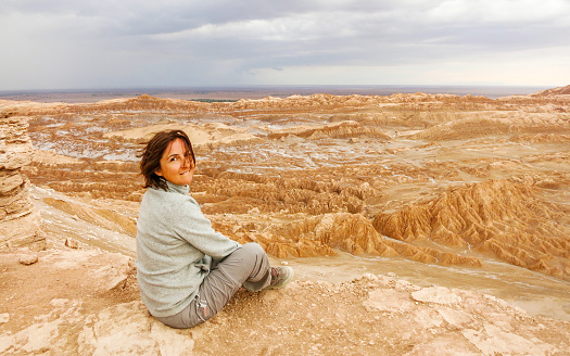 Portrait of woman sitting at cliff edge, looking at camera and admiring Moon Valley, Atacama Desert, Chile during sunset. Female traveler explores Valle de La Luna. Beautiful high angle view. Travel destination South America background with copy space.