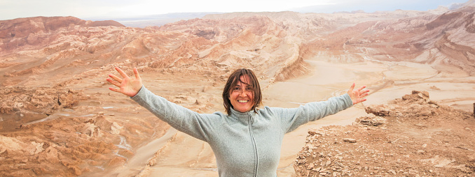 Portrait of a happy woman standing on the edge of a cliff with her arms wide open, looking at the camera in Valle de la Luna, Atacama Desert, Chile. Beautiful female traveller exploring Moon Valley, high angle view. Travel destination South America background with copy space.