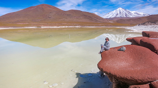 Young female white adult Caucasian sitting on red rocks Piedras rojas looking at camera over Salar de Talar. Snow-capped volcano Miniques reflected in turquoise lake. Idyllic Atacama Desert, volcanic landscape - San Pedro de Atacama