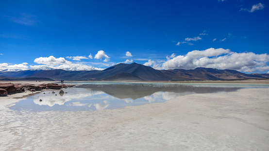 Blue sky reflection and unrecognisable tourists at the Tuyajto lagoon and salt lake in the Altiplano (high Andean plateau) over 4000 metres above sea level with salt crust on the shore, Antofagasta region, South America.