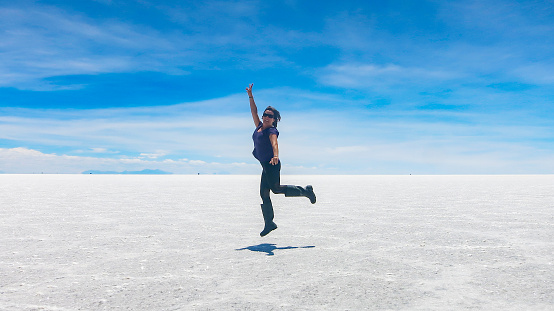 Woman with sunglasses jumping high with arms up and a smile on her face over the white salt flat lake Salar de Uyuni, blue sky. Endless boundless infinite vastness dreamy landscape in Potosi Bolivia South America. Travel destination background with copy space