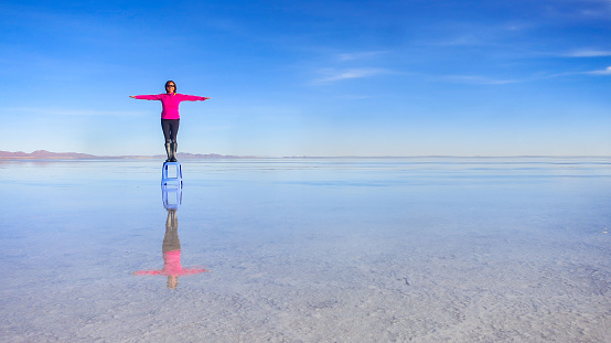 Woman with sunglasses standing on a chair with her arms wide open, like a cross and a smile on her face on the white salt flat lake in Salar de Uyuni, on a sunny day with blue sky. Endless boundless infinite vastness dreamy landscape in Potosi, South America. Travel destination background with copy space.