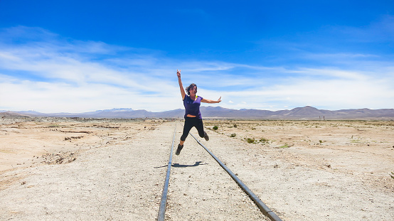 Woman jumps on railway tracks at the Bolivian border. Railway in the salt flats of Uyuni on the border between Bolivia and Chile. Blue sky on a sunny day. Travel destination background with copy space