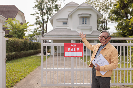 A friendly senior real estate agent smiles at the camera, confidently pointing to a house for sale.