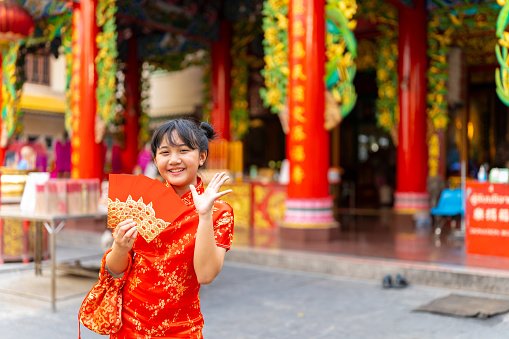 Chinese lunar new year festival and tradition holiday celebration concept. Portrait of Happy Little Asian girl in Chinese red dress holding money gift in red envelopes in front of in Chinese temple shrine.