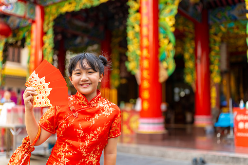Chinese lunar new year festival and tradition holiday celebration concept. Portrait of Happy Little Asian girl in Chinese red dress holding money gift in red envelopes in front of in Chinese temple shrine.