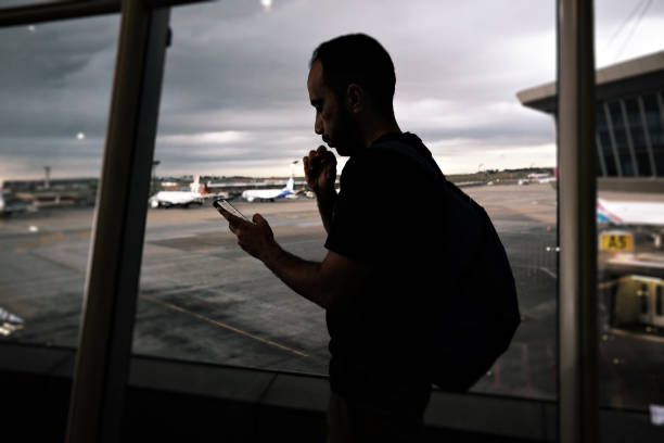 Backlit backpacker checking mobile phone while waiting for departure in front of the airport glass window.
