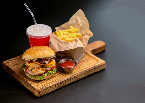 Classic burgers with becon, meat, cheese, onion, tomato and cheese and integral bun. On textured black board. Close up. Fried potatoes and glass of cola.