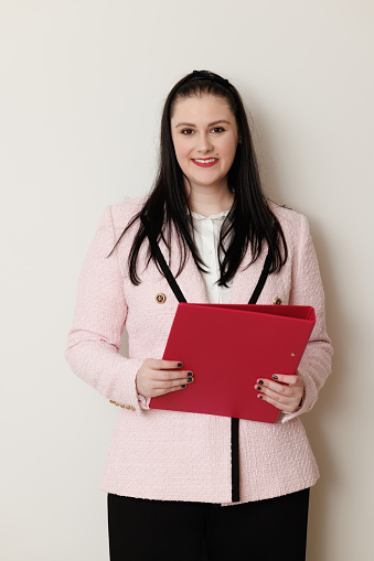 Smiling businesswoman holding a red folder in front of a plain wall.