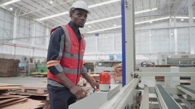 Young adult African engineer with protective workwear operating the cardboard processing machine using control panel at a paper factory. Focused male engineer controlling the production line at workplace.